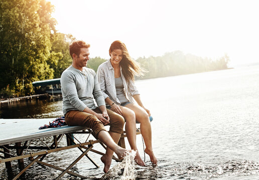 Vacation, couple and sitting on dock in nature for holiday, fun and splashing feet in water in summer. Happiness, people and romantic getaway by lake for adventure, commitment or love in relationship
