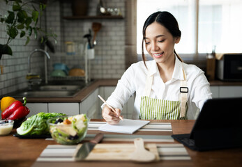 Young attractive asian woman in apron working from home, sitting in kitchen room and learning to cook online