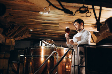 Young brewer wearing a leather apron is testing beer at a modern brewery