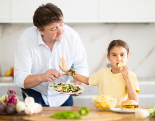 Little daughter eats chips in the kitchen, father offers her healthy vegetable salad