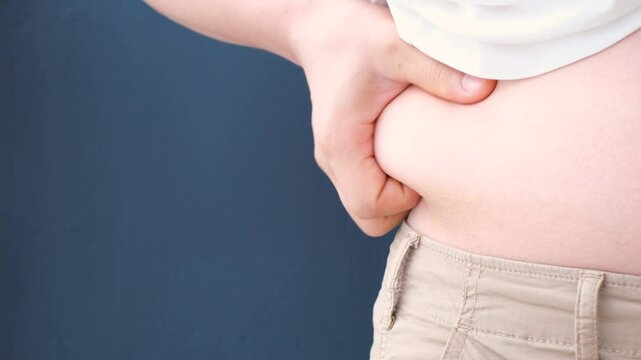 Male hand holding excessive belly fat on blue background, overweight concept.