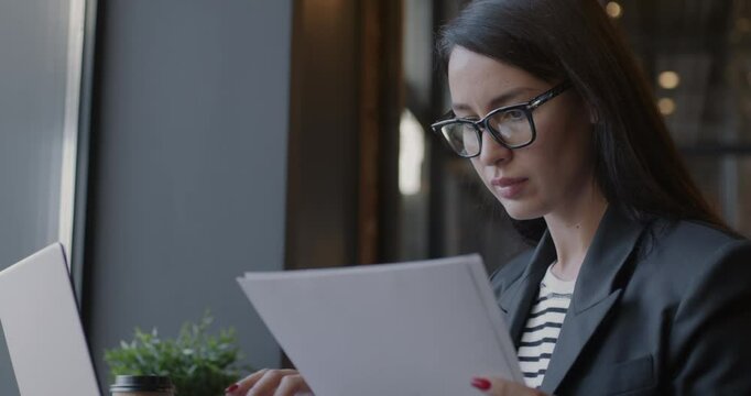 Female lawyer reading professional documents and working with laptop computer typing in cafe. Businesswoman and paperwork concept.