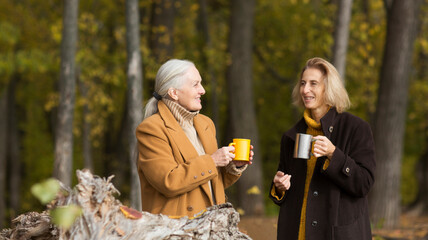 Two woman friends  drinks tea and enjoying a holiday  in autumn outdoor.  portrait of a smiling senior woman with an adult daughter relaxed outdoors 