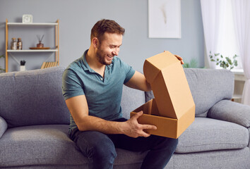 Happy man sits on the sofa at home living room, eagerly opening a box, parcel, or package from an online shopping order. Delighted by the contents, he revels in the joy of receiving delivery.