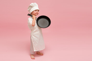 A funny child with a chef's hat and apron is posing with a cooking frying pan. Studio portrait on a pink backdrop. Kid aged 3 years (three year old boy)
