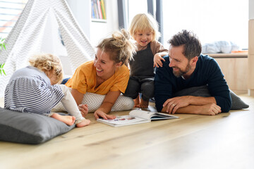 Young nuclear family playing with toys in a living room. Parents and children lying on floor, looking at children's story book, spending weekend day indoors.