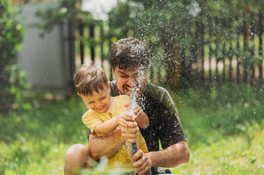 Happy man having fun with son splashing water from hose pipe in garden
