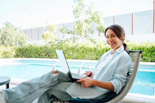Happy businesswoman using laptop sitting on lounge chair at poolside