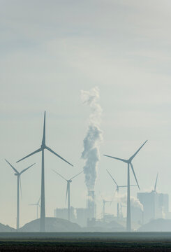Industrial chimney emitting smoke near wind turbines under sky