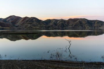 Before sunrise at the reservoir. Blue sky and mountains reflected in the water. Red cloud. Dry branches stick out from the water