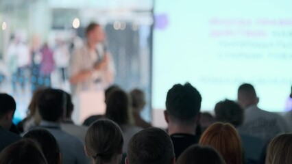 Diverse audience listening attentively at a business conference with a presenter speaking on stage.