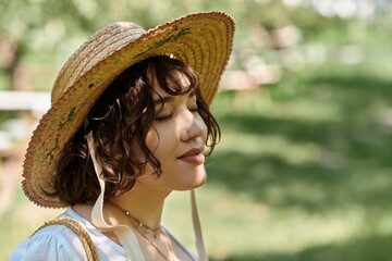 A young woman with brunette hair in a straw hat and white blouse relaxes in a sunny garden, eyes...