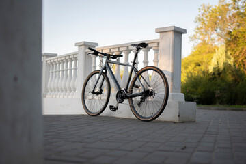 grey road bike is parked against a white wall. The bike has thin tires and is sitting on a concrete surface. The sun is shining brightly in the background