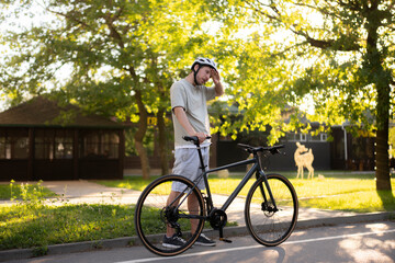 Fototapeta premium tired and exhausted man in a helmet takes a break from cycling in the park. He wears a grey shirt and white shorts, leaning on his black bike, surrounded by green grass and trees