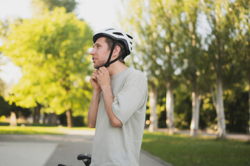 A man adjusts and puts on his white helmet while standing next to his bicycle in a park