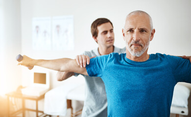 Obraz premium Rehabilitation, physical therapist and mature man with dumbbell in clinic for physiotherapy, support or mobility exercise. Lens flare, health worker and patient for help, muscle injury or recovery