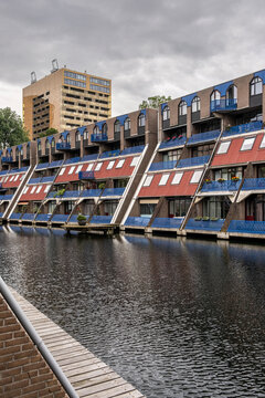 A captivating image of tilting modern brick apartments, featuring vibrant blue and red details, along a canal on a cloudy day, showcasing urban planning and residential architecture in Rotterdam