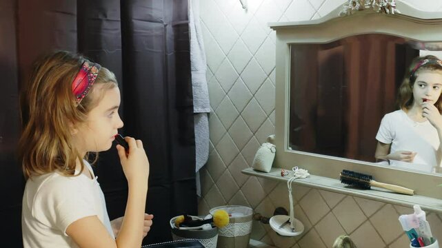 A cute little girl trying lipstick on her face for the first time, while in the bathroom in front of the mirror. Making a mistake, spreading too much color over her lips.