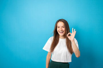 Teenager portrait on blue background, shows ok sign with fingers, young cute emotional student, smiling face with joyful emotion, fashionable curly hair