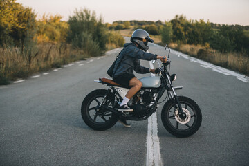 Obraz premium On the road, a biker drives his cafe racer motorcycle at sunset.