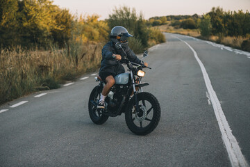 Obraz premium On the road, a biker drives his cafe racer motorcycle at sunset.