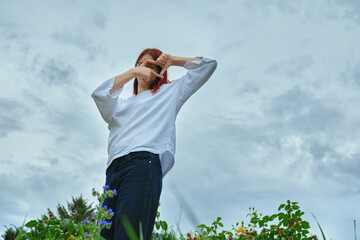 Young red-haired woman in a white shirt and glasses frames with her hands in a field of flowers under an overcast sky.