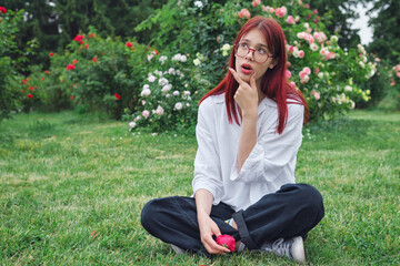 Teenage girl with red hair and glasses sitting in a blooming garden, pondering with a thoughtful expression, holding a rose