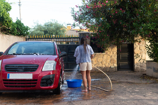 Back view of woman washing red car in driveway with garden hose