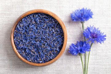 Dried blue cornflower petals in wooden bowl on linen fabric. Edible flowers of Centaurea cyanus, also known as bachelors button, flowers with intense blue pigment, used for tea and salad decorations.