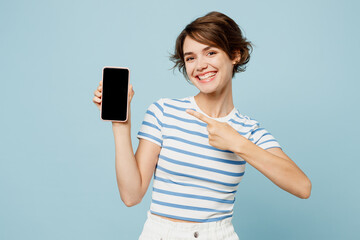 Naklejka premium Young woman wear striped t-shirt casual clothes hold in hand use mobile cell phone point index finger on blank screen workspace area isolated on plain pastel light blue background. Lifestyle concept.