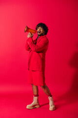 A young Indian man in a vibrant red suit holding a red megaphone.