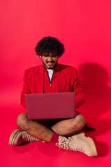 A young Indian man in vibrant attire sitting on the floor, engrossed in his laptop.