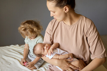 Older Caucasian sister sitting on bed in room with little sibling and mommy beautiful mother holding baby in hands eldest daughter jealous of her mother's newborn baby