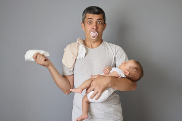 Shocked astonished busy father with diaper in hands and dirty clothing on shoulders holding newborn...