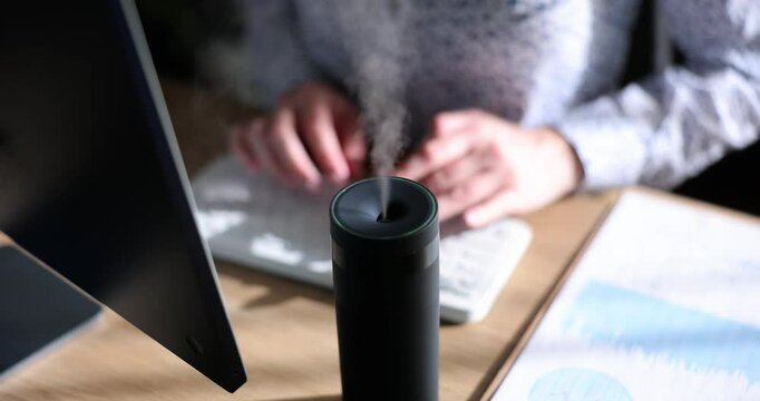 Freelancer types article on keyboard inhaling scent of essential oils. Modern diffuser spreads aroma and humidifies air in room slow motion
