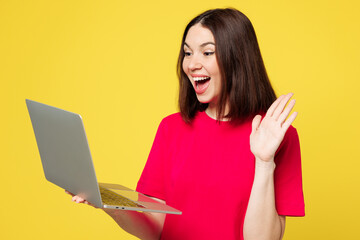 Young surprised fun IT woman wear pink t-shirt casual clothes hold use work on laptop pc computer waving hand talk speak isolated on plain yellow orange background studio portrait. Lifestyle concept.