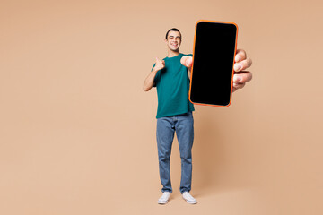 Full body young middle eastern man wear blue t-shirt casual clothes hold use blank screen workspace area mobile cell phone show thumb up isolated on plain pastel beige background. Lifestyle concept.