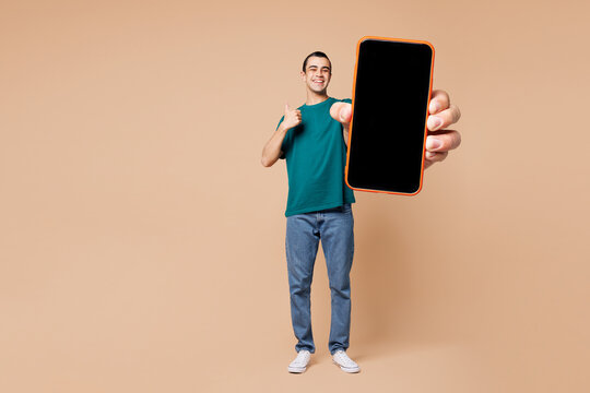 Full body young middle eastern man wear blue t-shirt casual clothes hold use blank screen workspace area mobile cell phone show thumb up isolated on plain pastel beige background. Lifestyle concept.
