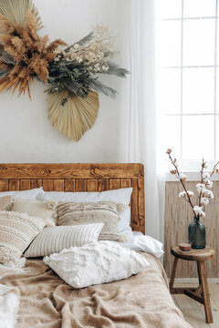 Cozy rustic bedroom with boho ethnic decor. Large windows. Double wooden bed with many pillows and handmade textiles. Wooden furniture. Wooden stool. Nobody. Large windows in appartment