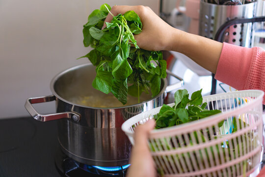 A woman is blanching or cooking spinach in a pot on a stove. She is holding a basket of spinach in her hand, home cooking concept