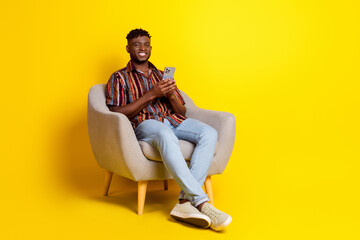 Full size photo of nice young man sit armchair smart phone wear shirt isolated on yellow color background