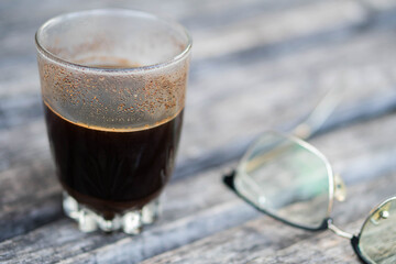 Glass of black coffee with eyeglasses on wooden surface