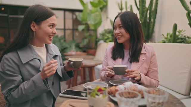 Two diverse women enjoy a conversation and coffee while sharing breakfast at a cozy cafe, exhibiting friendship and enjoyment.