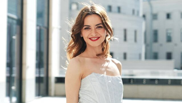 Young beautiful smiling brunette female in trendy evening silver dress. Sexy carefree woman posing in the street at sunny day. Fashionable model with evening bright makeup and red lips, outdoors