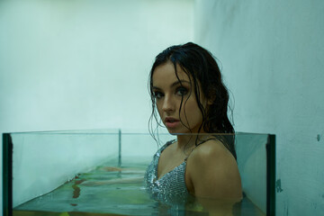 A beautiful young woman with wet hair, wearing a shimmering top, emerges from a glass tank filled with water.