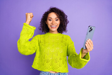Photo of curly american young girl in green sweater using smartphone fist up winning playing lottery isolated on purple color background