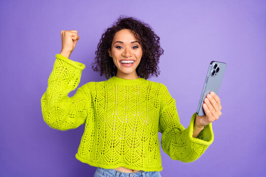 Photo of curly american young girl in green sweater using smartphone fist up winning playing lottery isolated on purple color background