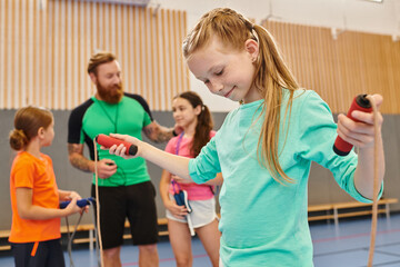 A diverse group of kids engages in an energetic gym session, with a girl confidently holding a jump rope as the male teacher instructs and guides them. © LIGHTFIELD STUDIOS