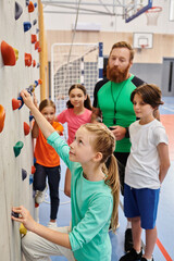 A man teacher instructs a diverse group of kids and adults as they stand around a climbing wall, preparing to embark on an adventurous climbing challenge. 