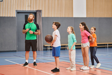 A man stands before a group of kids, holding a basketball and providing guidance in a vibrant, engaged classroom setting.